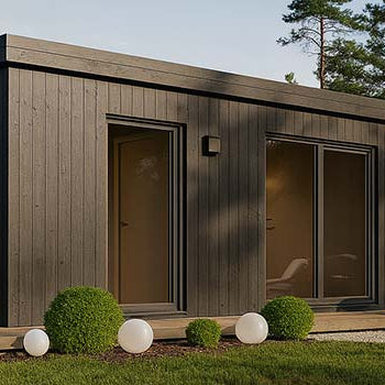 Sleek black timber garden room with patio loungers nestled among pine trees at sunset.