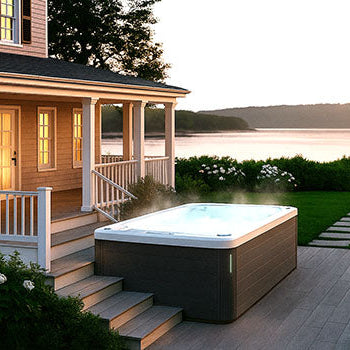 Steaming hot tub on lakeside deck at sunset beside a classic home with glowing windows.