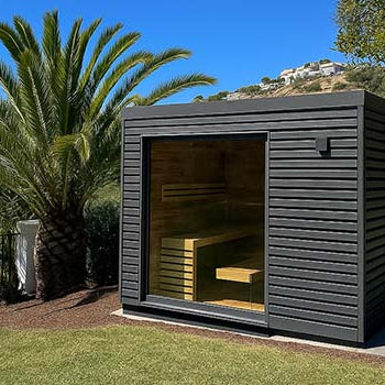 Contemporary black outdoor sauna with glass front on a sunny lawn with palm trees and hillside views.