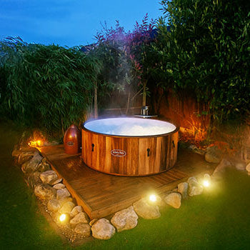 Steaming round wooden hot tub on deck with garden lighting at dusk.