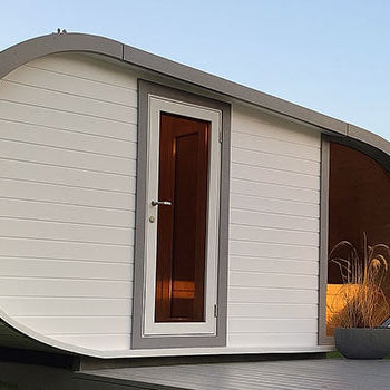 Modern white barrel sauna with curved roof on wooden deck in grassy garden at sunset.