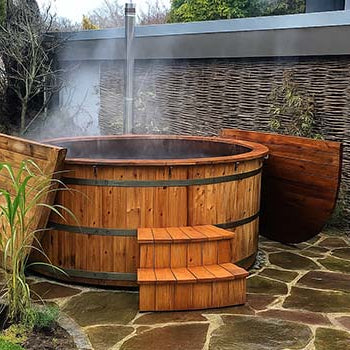 Round wooden hot tub with steam rising on a flagstone patio beside woven fencing and garden greenery.