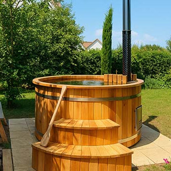 Traditional wooden hot tub with chimney in a sunny garden surrounded by trees and greenery.
