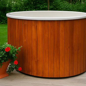 Round wooden hot tub on outdoor deck with flower planter and green garden background.