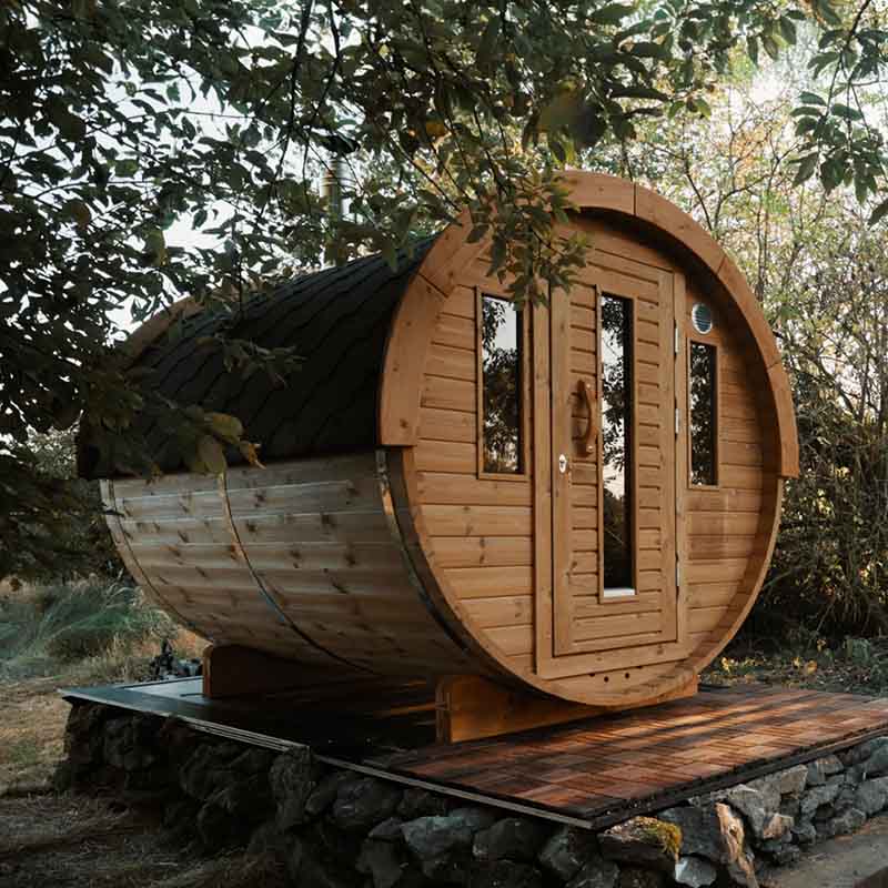 Outdoor barrel sauna with a red shingle roof and wooden cladding, set on a raised deck surrounded by forest landscaping.