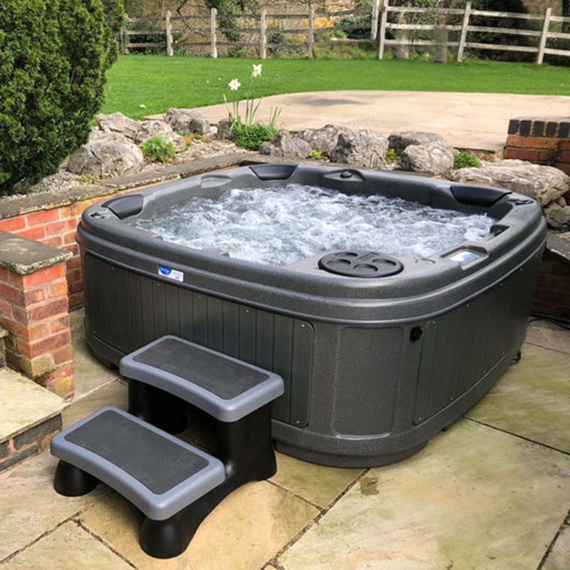 Dark grey rotomoulded hot tub with bubbling water and matching steps, placed on a stone patio in a landscaped garden.