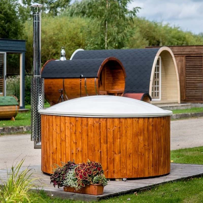 Round wooden hot tub with white insulated cover and stainless steel flue, set outdoors on a timber deck with barrel saunas in the background.