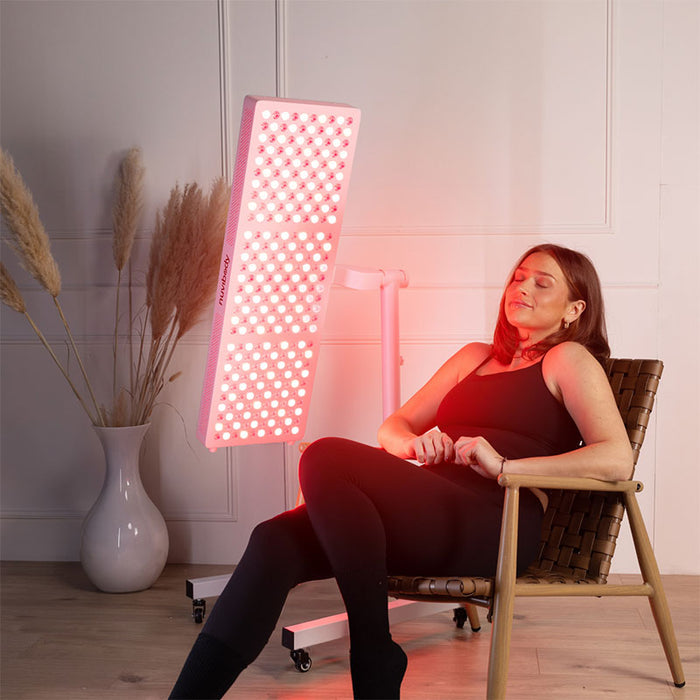 Woman enjoying a relaxing red light therapy session with the Nuvibody LITE 1500 panel mounted on an adjustable stand, ideal for skin rejuvenation and half-body treatment at home.
