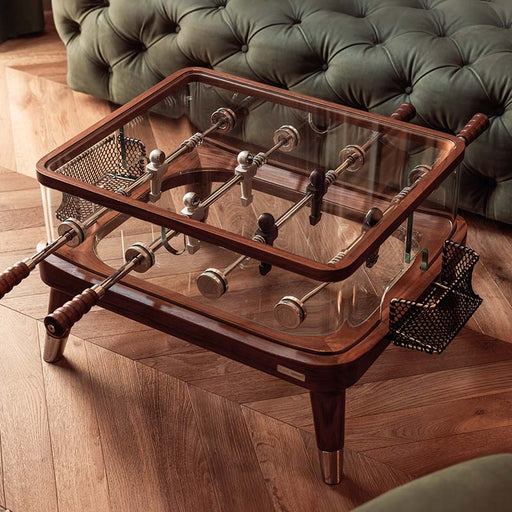 Luxury Teckell Intervallo foosball coffee table with crystal glass sides, solid wood frame, and stainless steel rods, styled in a sophisticated lounge with parquet flooring.