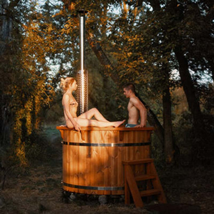 A couple relaxing in a 1.9m thermowood hot tub with a chimney, set in a peaceful woodland area.