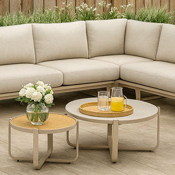 beige modern L-shaped garden sofa with bolster cushions and round nesting tables on tiled patio with wooden fence and olive trees.