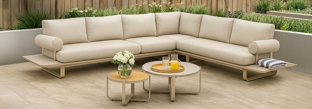beige modern L-shaped garden sofa with bolster cushions and round nesting tables on tiled patio with wooden fence and olive trees.