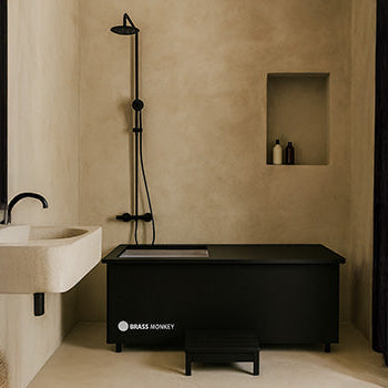 Matte black Brass Monkey ice bath with steps in a minimalist beige bathroom with walk-in shower and floating sink.