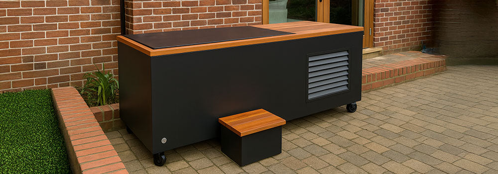 Modern black ice bath with wood top and wheels on a stone patio beside red brick house.