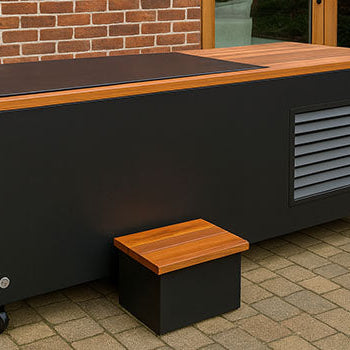 Modern black ice bath with wood top and wheels on a stone patio beside red brick house.