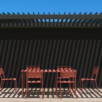 Black pergola casting striped shadows over terracotta dining set on wooden deck with stone wall and tropical plants.