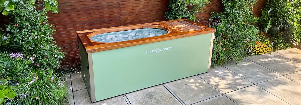 Mint green Brass Monkey ice bath filled with ice on a patio surrounded by greenery and wooden fencing.