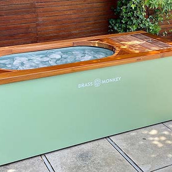 Mint green Brass Monkey ice bath filled with ice on a patio surrounded by greenery and wooden fencing.