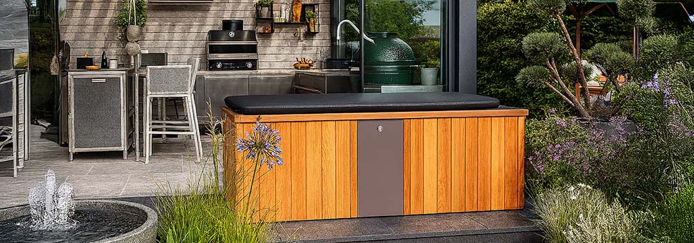 Cedar-clad ice bath with black cover in a modern garden kitchen patio surrounded by lush plants and water feature.