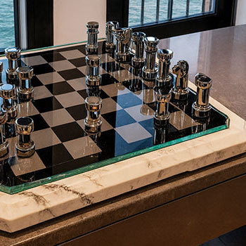 Modern chrome chess set with black glass board on marble base by large windows overlooking water.