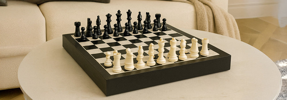 Classic black and white chess set on a marble coffee table in a neutral-toned living room with plush seating and a vintage mirror.