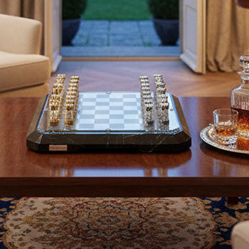 Sophisticated luxury chess board with gold and silver pieces on a black marble base, set atop a polished wood coffee table in a refined lounge with garden view.