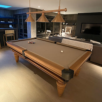 rustic wood pool table with gray felt in a bright farmhouse-style game room with large windows and industrial lighting.