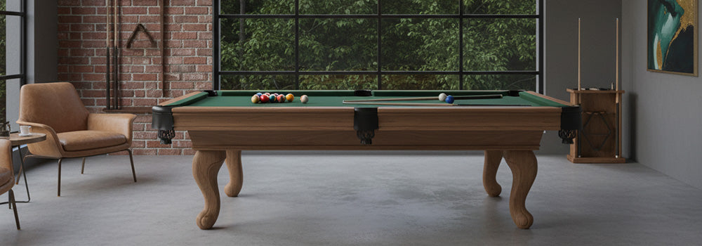 Classic wooden pool table with traditional carved legs in an industrial-style games room with exposed brick walls and large black-framed windows.