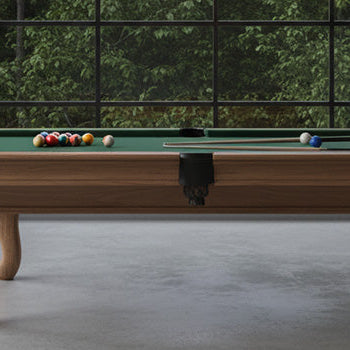 Classic wooden pool table with traditional carved legs in an industrial-style games room with exposed brick walls and large black-framed windows.