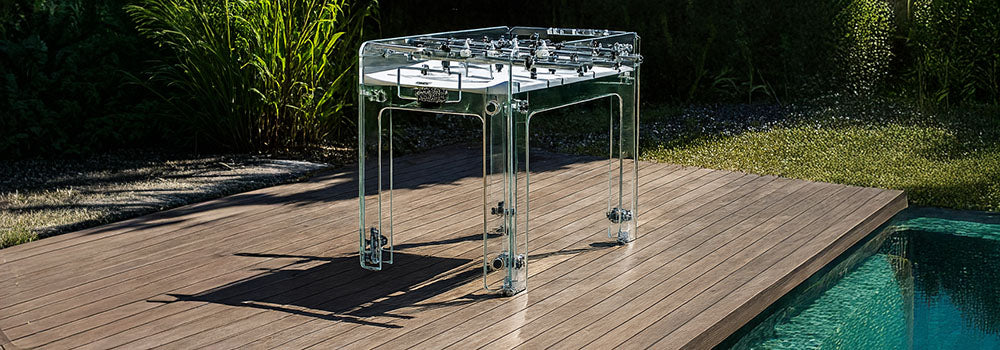 Transparent foosball table on a wooden deck beside a turquoise pool surrounded by lush greenery.