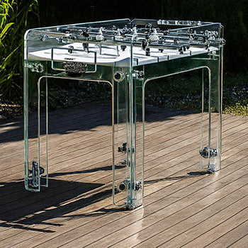 Transparent foosball table on a wooden deck beside a turquoise pool surrounded by lush greenery.
