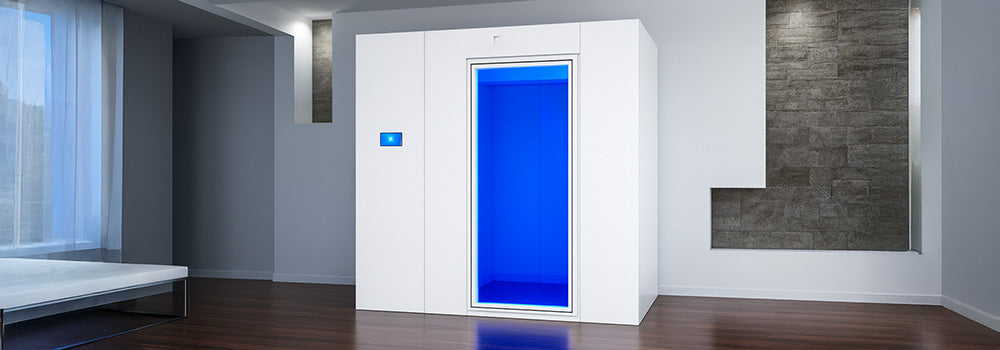 Modern white cryotherapy chamber with blue-lit interior in a minimalist room with dark wood floors and stone accent wall.