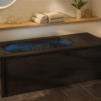 Dark wood ice bath in tranquil spa room with brick wall, potted plant, and soft natural lighting.