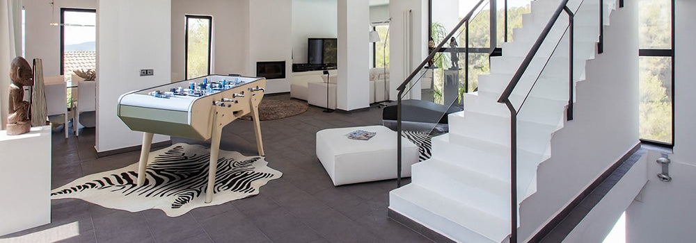 Designer foosball table in a bright, modern open-plan living room with white walls, glass railings, and zebra print rug.