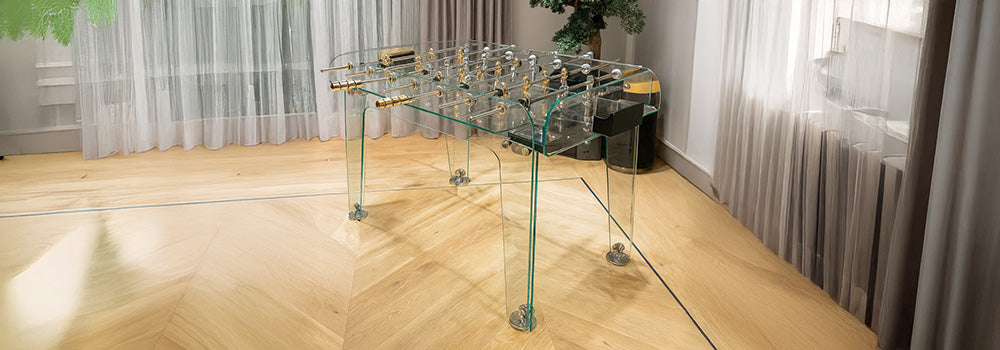 transparent glass foosball table with gold accents in modern interior with herringbone floor and bonsai tree.