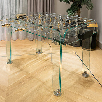 transparent glass foosball table with gold accents in modern interior with herringbone floor and bonsai tree.