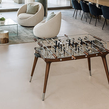 luxury glass foosball table with walnut legs in a bright open-plan living and dining area with modern furniture and large windows overlooking a terrace.