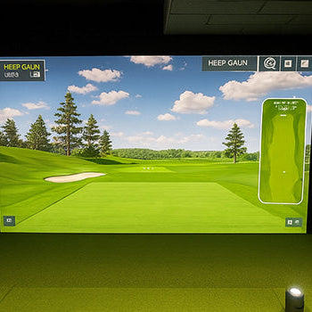 Indoor golf simulator room with green turf, dual screens, wall padding, and complete golf club set.