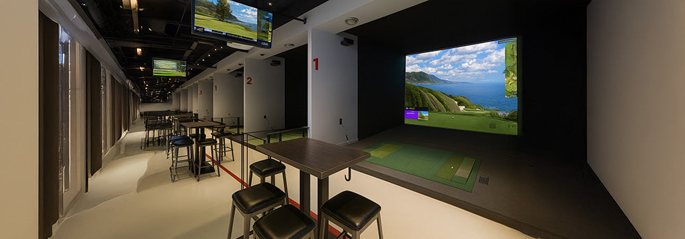 Upscale indoor golf simulator lounge with numbered bays, ocean course display, and high-top seating.