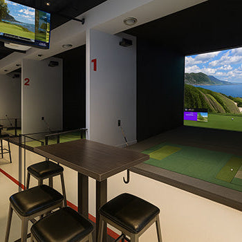 Upscale indoor golf simulator lounge with numbered bays, ocean course display, and high-top seating.