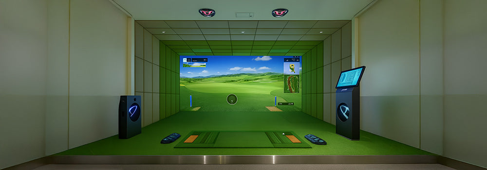 Indoor golf simulator setup with turf mat, fairway projection, and touchscreen kiosk in a softly lit room.