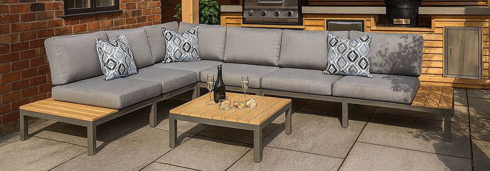 Grey garden corner sofa with wood accents and matching table on a stone patio beside a modern outdoor kitchen.