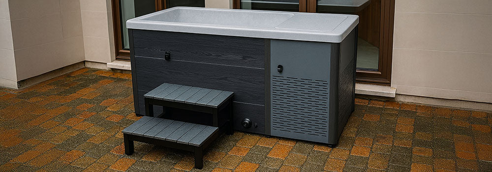 Modern grey ice bath with steps on a wet cobblestone patio beside a stone accent wall and glass doors.