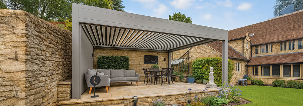 Sleek grey pergola over sandstone patio with dining and lounge furniture beside traditional stone cottage.
