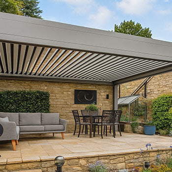 Sleek grey pergola over sandstone patio with dining and lounge furniture beside traditional stone cottage.
