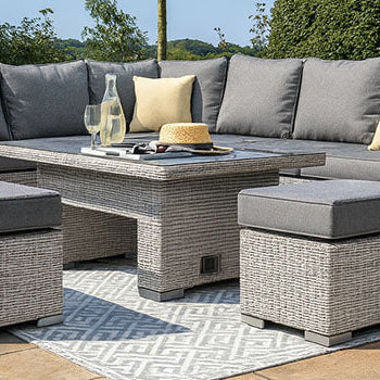 Grey rattan corner sofa set with yellow cushions and matching table on a tiled patio with rug and landscaped garden.