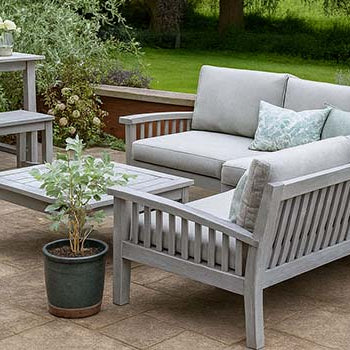 grey wooden garden sofa set with cushions and matching dining table on a tiled patio surrounded by lush countryside greenery.