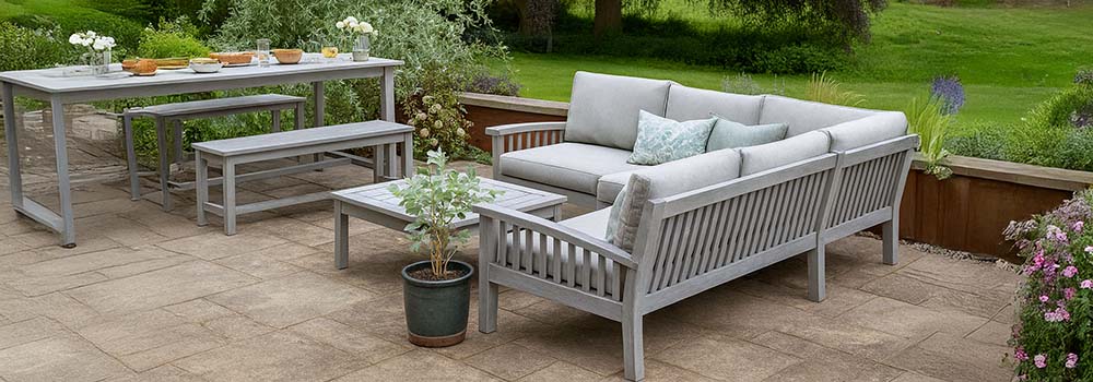 grey wooden garden sofa set with cushions and matching dining table on a tiled patio surrounded by lush countryside greenery.