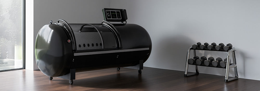 Hard-shell hyperbaric oxygen chamber in a modern fitness room with dark wood flooring, a dumbbell rack, and floor-to-ceiling windows.