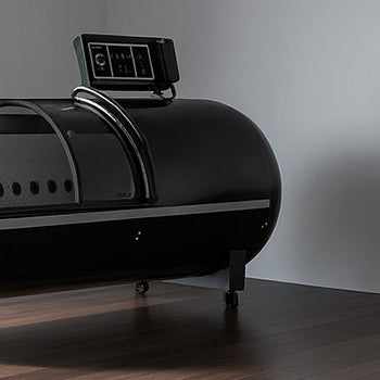 Hard-shell hyperbaric oxygen chamber in a modern fitness room with dark wood flooring, a dumbbell rack, and floor-to-ceiling windows.